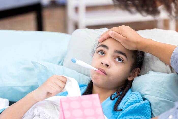 African American mother checks her daughter's temperature. The girl has a thermometer in her mouth and is holding a box of tissues.