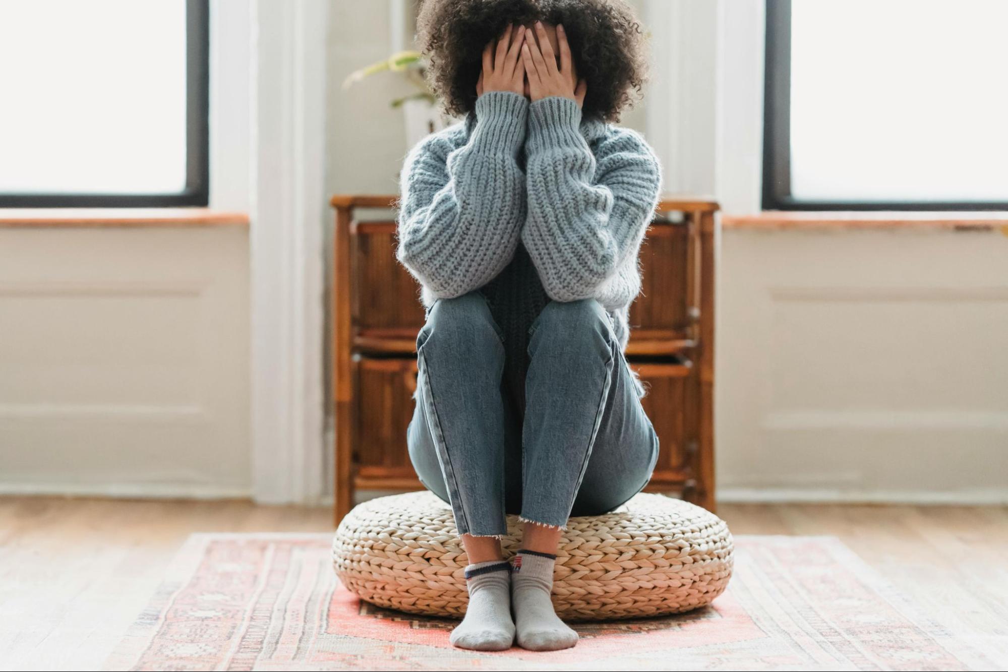 young-person-covering-face Young person covering face with hands, sitting on a round cushion in a bright room